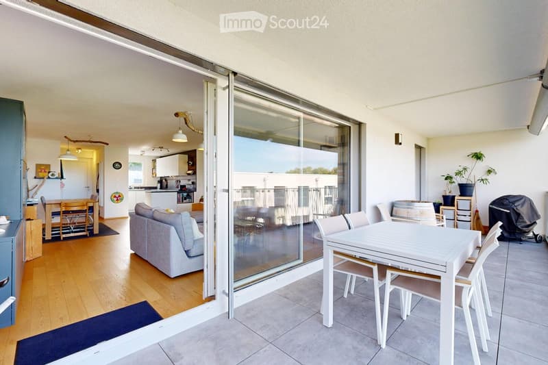 Balcony with a white table and chairs, a sofa visible through the sliding glass door, and indoor dining area with a table and chairs.