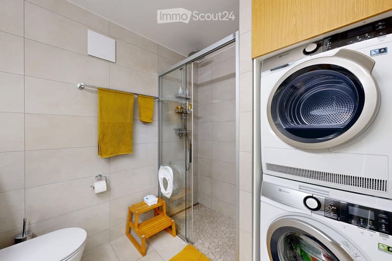 Modern bathroom with a toilet, shower, washing machine, and dryer. Features include a wooden stool, yellow towels, and a glass shower enclosure.