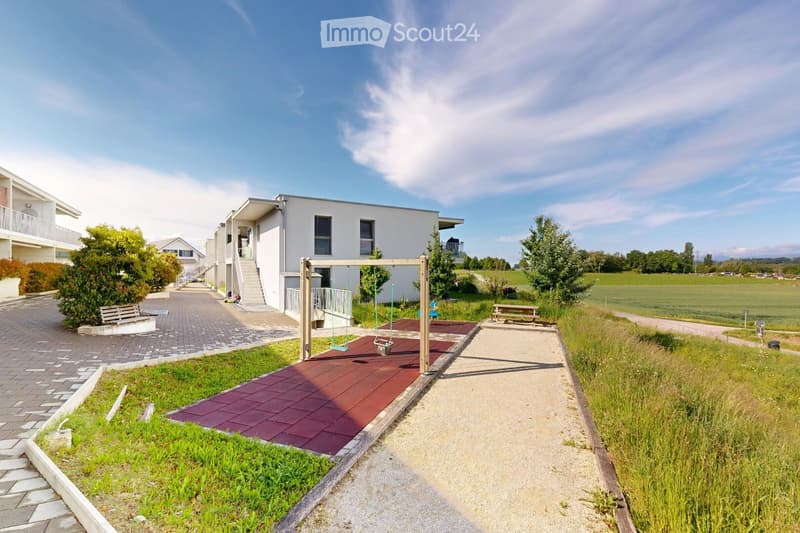 Outdoor area with a playground, swings, benches, paved walkways, and modern residential buildings in the background.