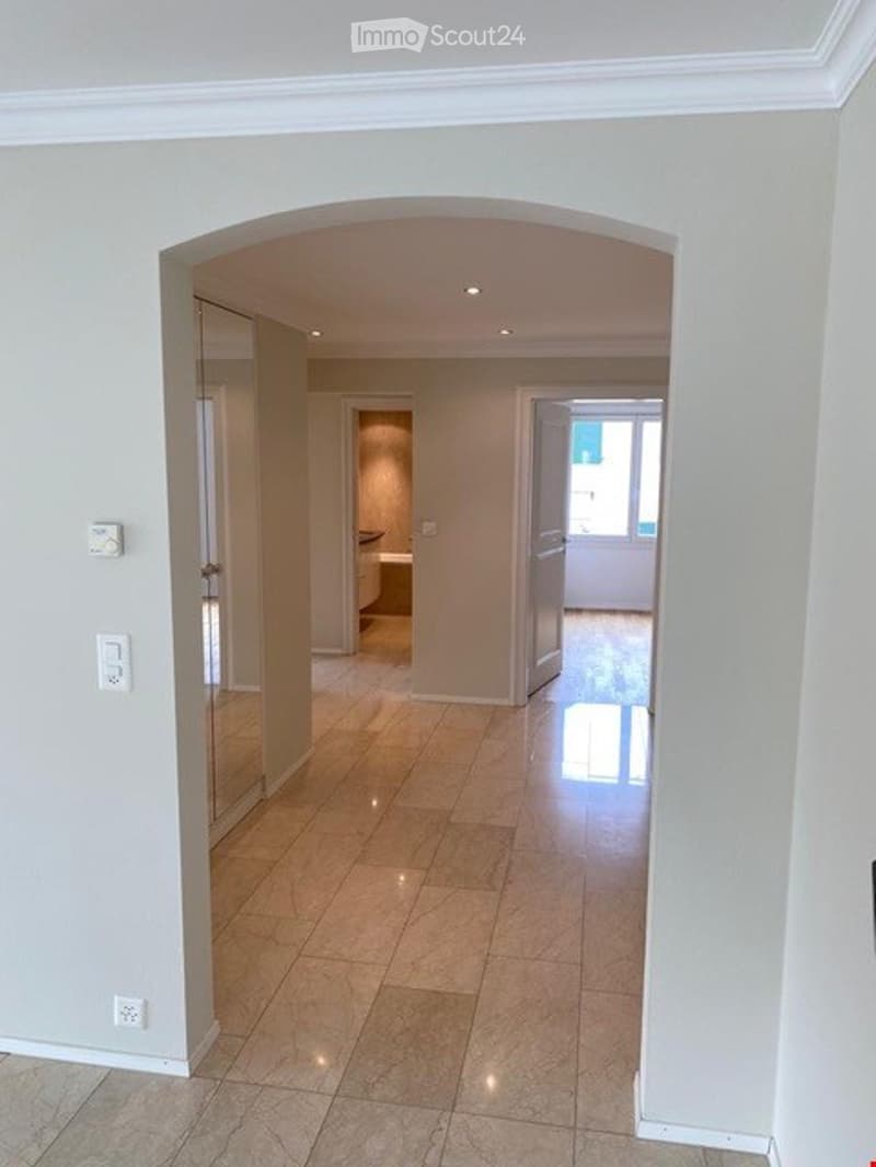 Bright hallway with beige tiled flooring, white walls, and recessed lighting. An open doorway leads to a bathroom with a visible bathtub and a windowed door to the outside.