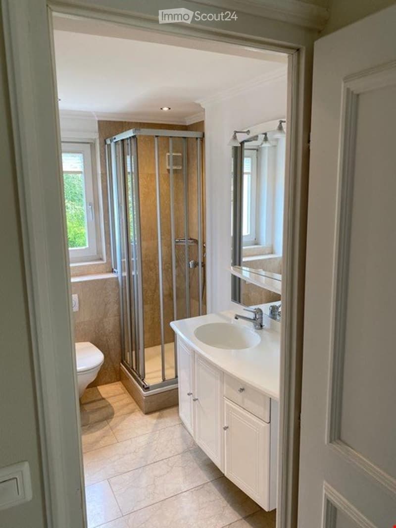 Modern bathroom with a white sink, mirror, and a glass-enclosed shower stall. The space features light-colored tiles and a window providing natural light.