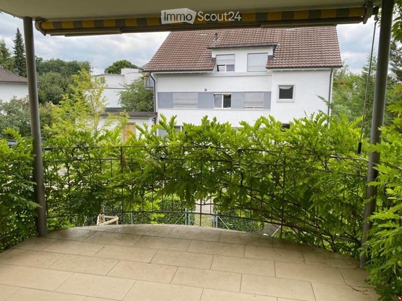Balcony with tiled floor, metal railing, and lush greenery, overlooking a residential area with houses and trees.