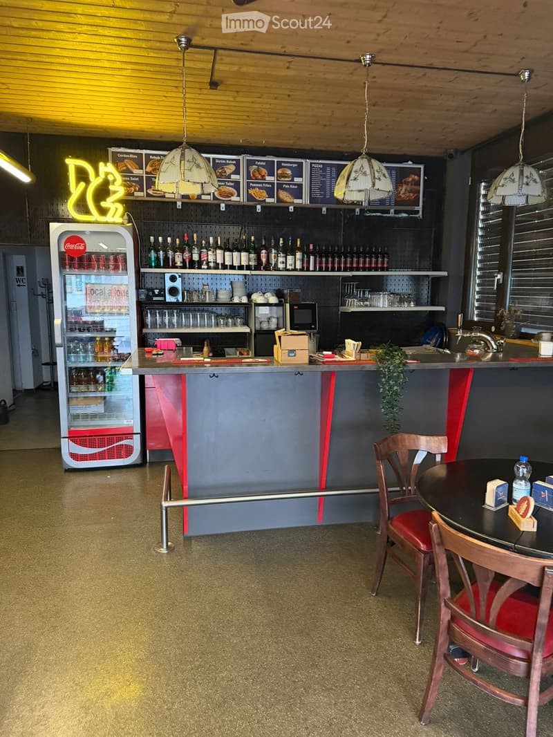 Wooden ceiling, modern decor, open kitchen with shelves, a Coca-Cola fridge, a large counter, red chairs, tables, and stools.