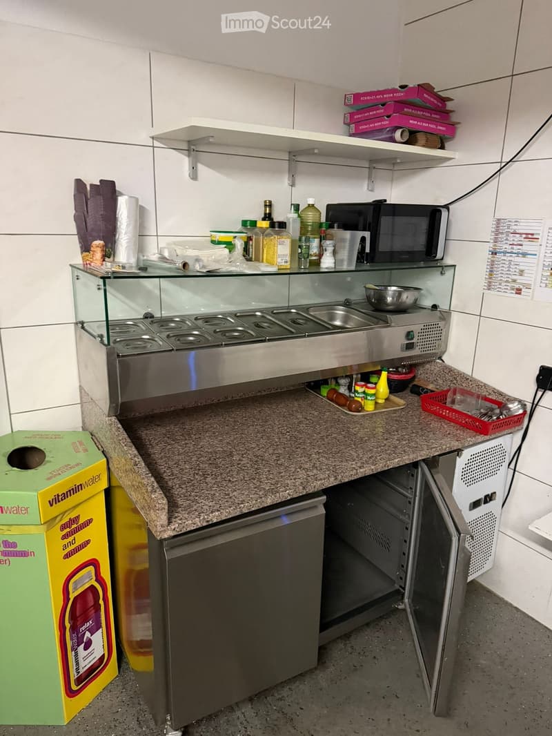 a kitchen with stainless steel countertop, a glass display case, shelves, bottles, containers, a microwave, and a refrigerator.