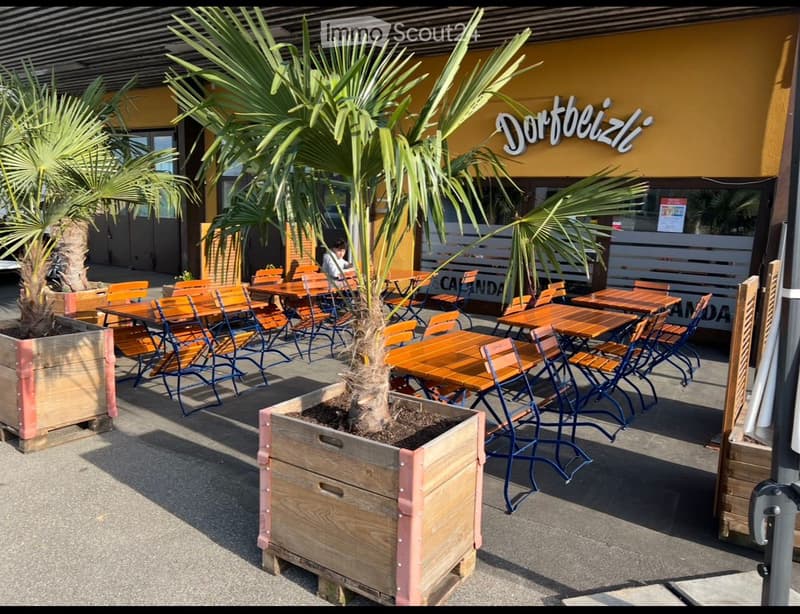 Outdoor seating area with wooden tables and chairs arranged in rows, featuring multiple potted palm trees on the ground, yellow building, signage with the text 'Dortbeizl', and an outdoor seating arrangement for customers.