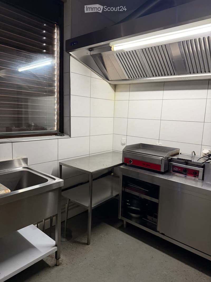 Commercial kitchen with stainless steel appliances, including a sink, stove, grill, and a worktable.