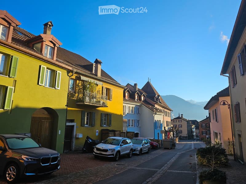 A picturesque European town street with colorful buildings, parked cars, and a clear blue sky. The architecture features multi-story houses with varied facades, shutters, and balconies. The street is paved with cobblestones, and mountains are visible in t