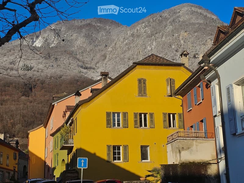 A vibrant yellow building with multiple windows and shutters, situated in a picturesque village with colorful houses and a mountain backdrop.