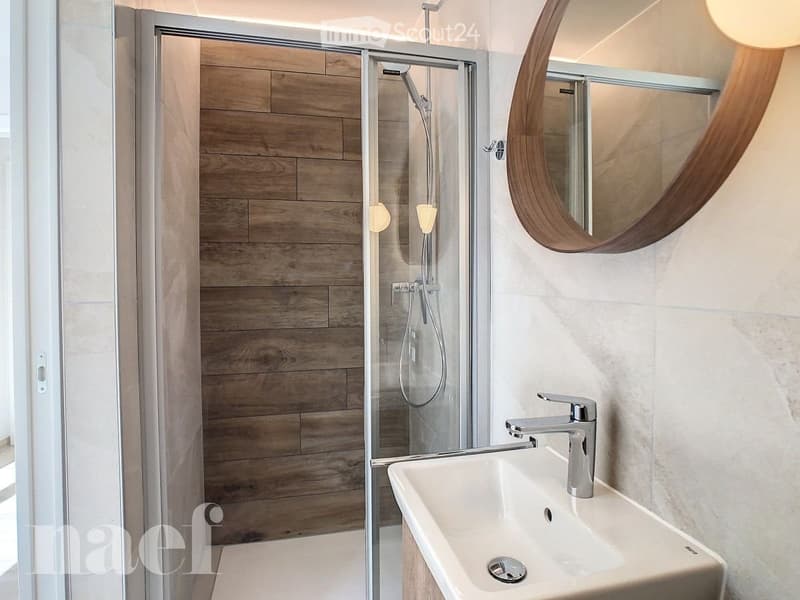 Modern bathroom with a white sink, chrome faucet, round wooden-framed mirror, and a glass-enclosed shower with wooden walls.