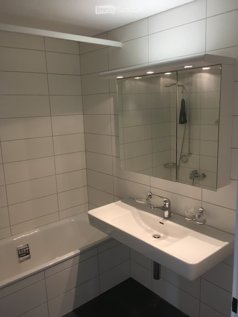 Modern bathroom with a white sink, chrome faucet, bathtub, and large mirror with built-in lighting. The walls are tiled in light gray.