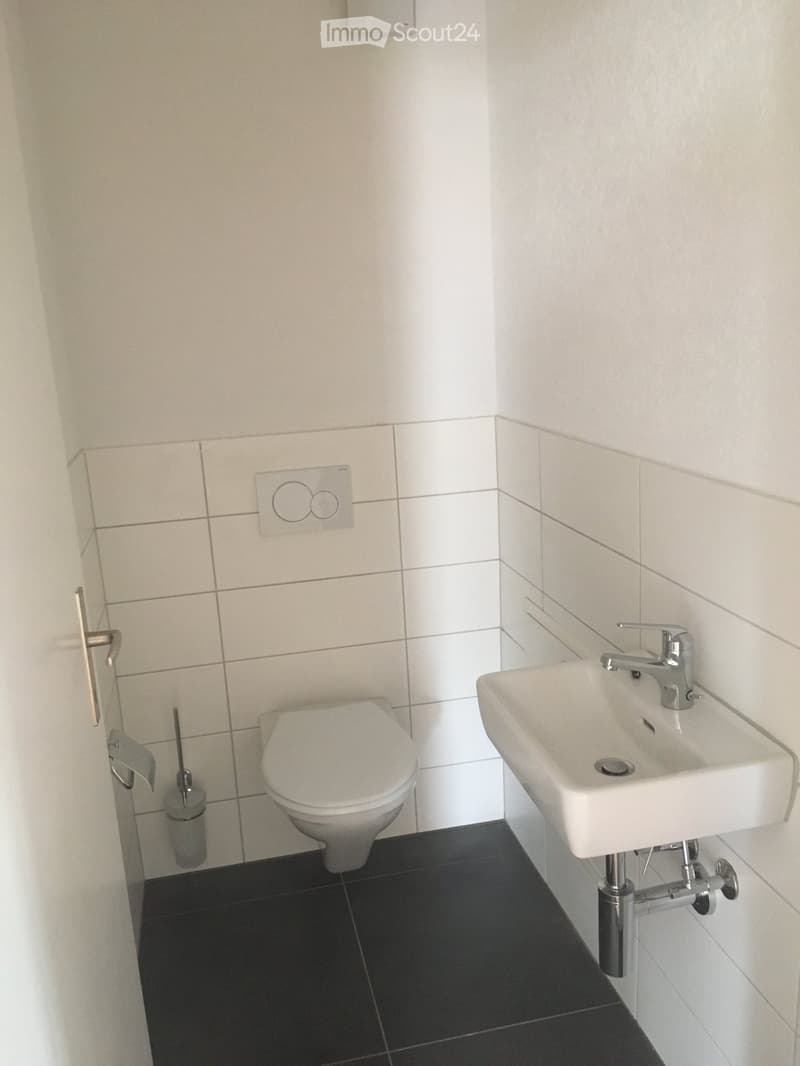 Modern bathroom with white tiled walls, black tiled floor, toilet, sink with chrome faucet, and toilet brush holder.
