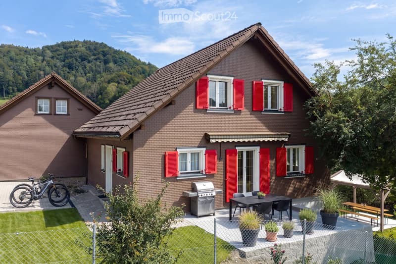 Two-story brick house with red shutters, surrounded by a well-maintained garden and outdoor seating area. Features include a barbecue grill, patio furniture, and bicycles parked nearby.