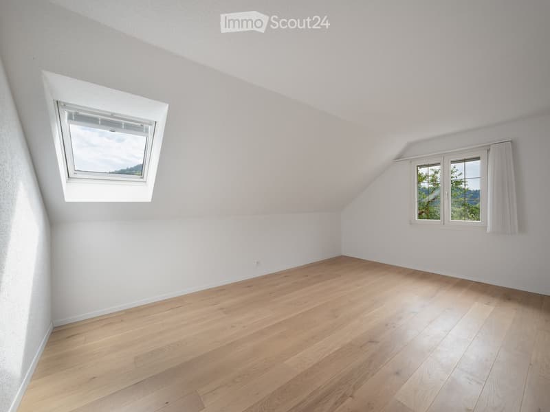 Empty attic room with wooden flooring, white walls, and two windows, one of which is a skylight.