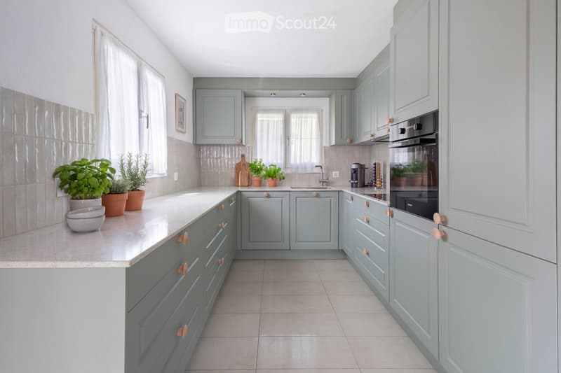 Modern kitchen with light blue cabinets, white countertops, and stainless steel appliances. Features a central island, built-in oven, and various kitchen items including potted plants and a coffee maker.