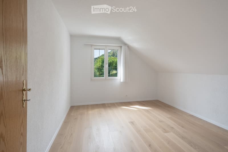 Empty attic room with wooden flooring, white walls, and a window with white curtains showing a green outdoor view.