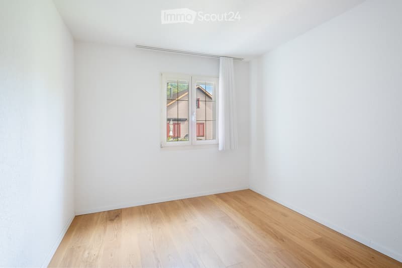 Empty room with light wooden flooring and white walls, featuring a large window with white curtains and a view of a neighboring house.