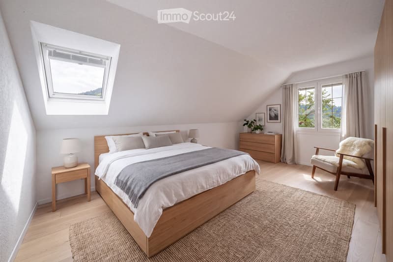 Cozy attic bedroom with wooden bed, nightstands, lamps, a plush chair, and a dresser with a plant and picture frames. Large windows and skylight provide natural light.