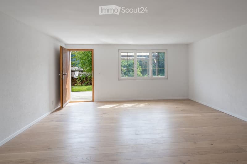 Empty room with light wooden flooring, white walls, and a large window with a view of greenery. A wooden door leads to an outdoor area.