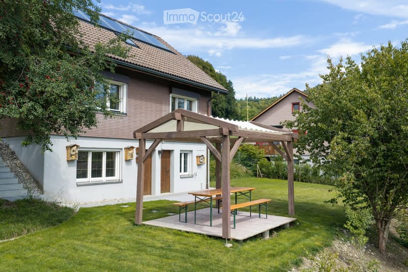 A well-maintained garden with a wooden pergola, picnic tables, and benches, surrounded by a two-story house with solar panels on the roof and a raised ground floor entrance.