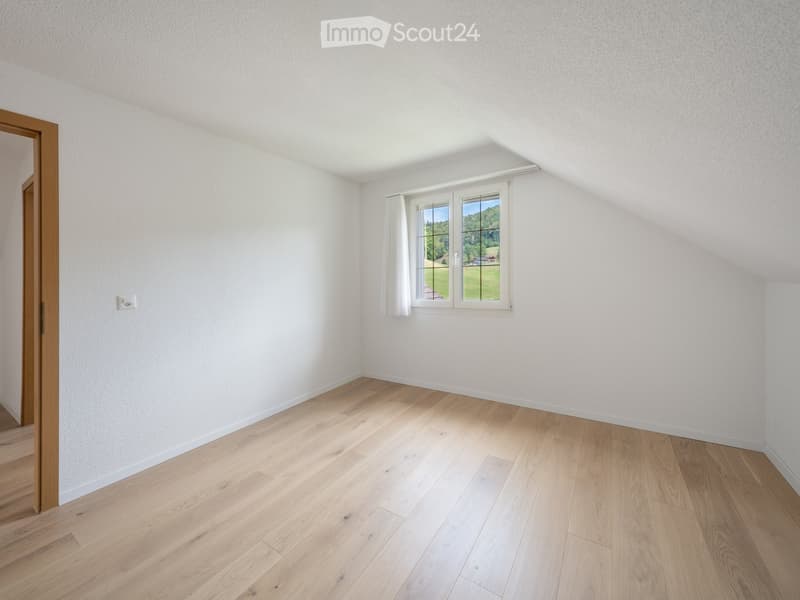 Empty room with light wooden flooring, white walls, and a window with white curtains. The room has a slanted ceiling typical of an attic space.