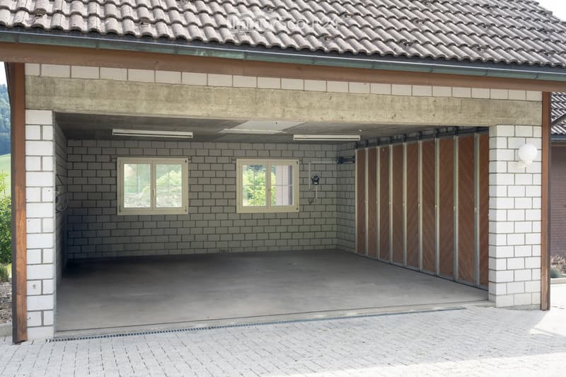 Garage with concrete walls, tiled floor, and wooden doors, featuring two windows and overhead lighting.