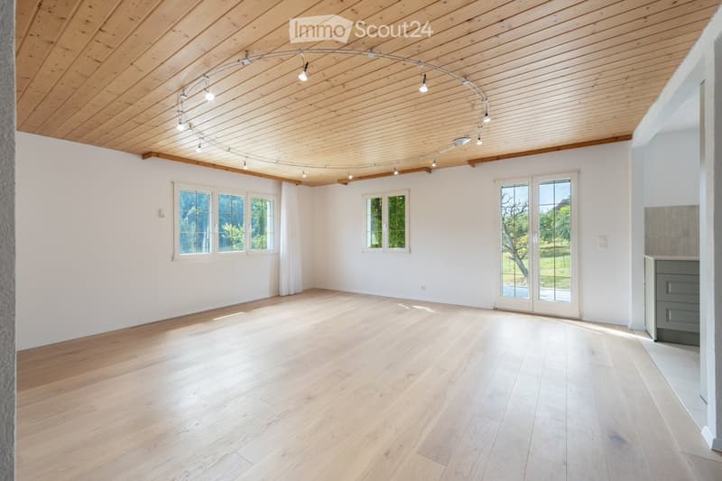Bright, empty room with wooden flooring and white walls, featuring large windows and a sliding glass door leading outside.