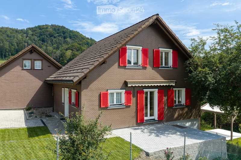 Two-story brick house with red shutters, white-framed windows, and a sloped roof. Features a covered entrance, a small garden, and a paved driveway.