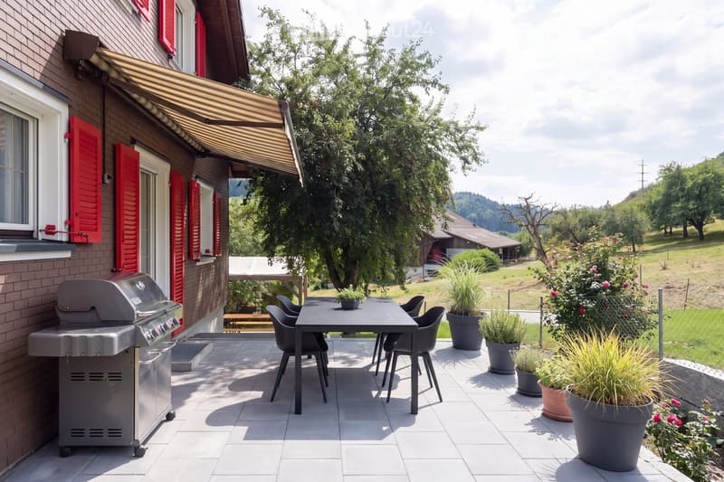 Outdoor patio with a grill, dining table, chairs, and potted plants, attached to a house with red shutters and a striped awning.