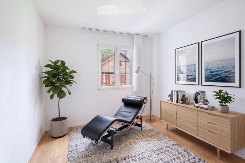 Modern, minimalist room with a black leather lounge chair, wooden sideboard with books and decorative items, large potted plant, framed artwork, and a floor lamp near the window.