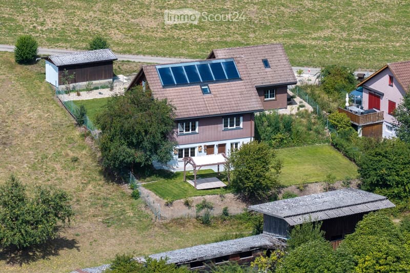 Aerial view of a residential area with multiple houses, including a central house with a solar panel roof, surrounded by greenery and smaller structures.