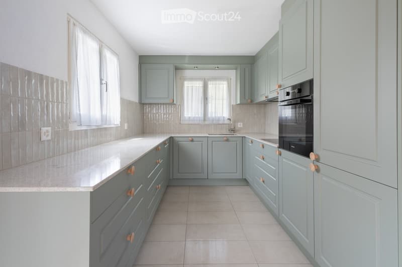 Modern kitchen with light blue cabinets, white countertops, and a built-in oven. Features tiled backsplash and large windows with white curtains.