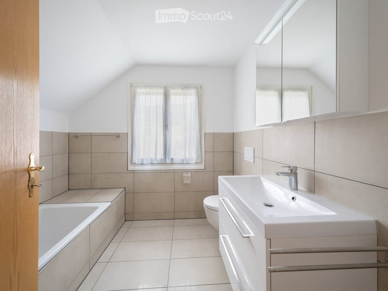 Modern bathroom with a bathtub, sink, toilet, and large mirror. Light-colored tiles and wooden door frame.