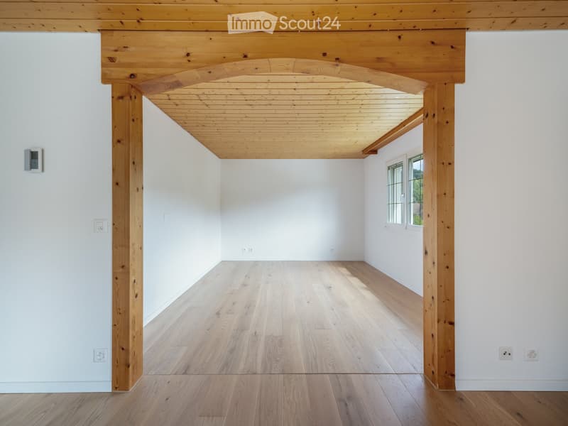 Empty room with light wooden flooring, white walls, and exposed wooden beams supporting a raised ceiling. A single window allows natural light to enter.