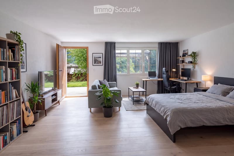 Bright, modern bedroom with a large bed, sofa, desk with laptop, and a bookshelf. Features large windows with curtains, a small table, and several potted plants.