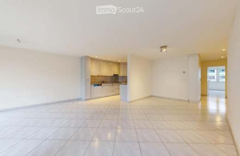 Empty kitchen and living area with white tiled flooring, white walls, and built-in kitchen cabinets.