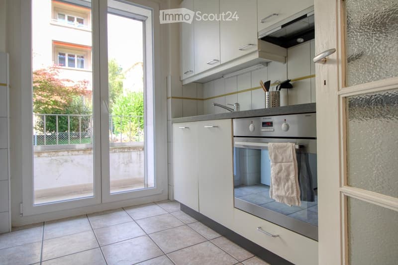 Modern kitchen with white cabinets, stainless steel oven, tiled floor, and a towel hanging on the oven door. A large window with a view of a balcony and greenery is visible.