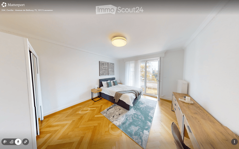 Bright bedroom with wooden flooring, a large bed with pillows and a blanket, a nightstand with a lamp, a small table, a rug, and a balcony with a view.