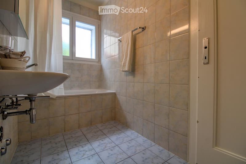 Bathroom with a sink, bathtub, tiled walls, and a window with a curtain. A towel hangs on a rack, and a small basket is placed on the sink counter.
