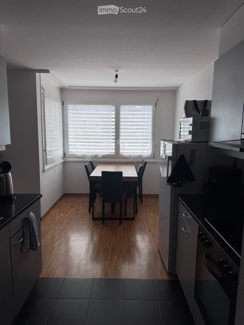 A modern kitchen and dining area with wooden flooring, large windows with blinds, a dining table with chairs, and kitchen appliances including a refrigerator, microwave, and oven.