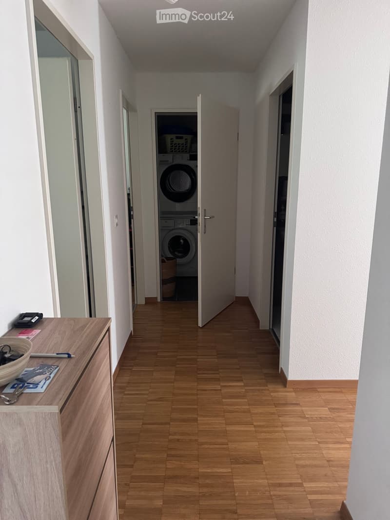 A narrow hallway with wooden flooring, white walls, and a small wooden cabinet on the left side. An open door leads to a laundry area with a washing machine and dryer visible.
