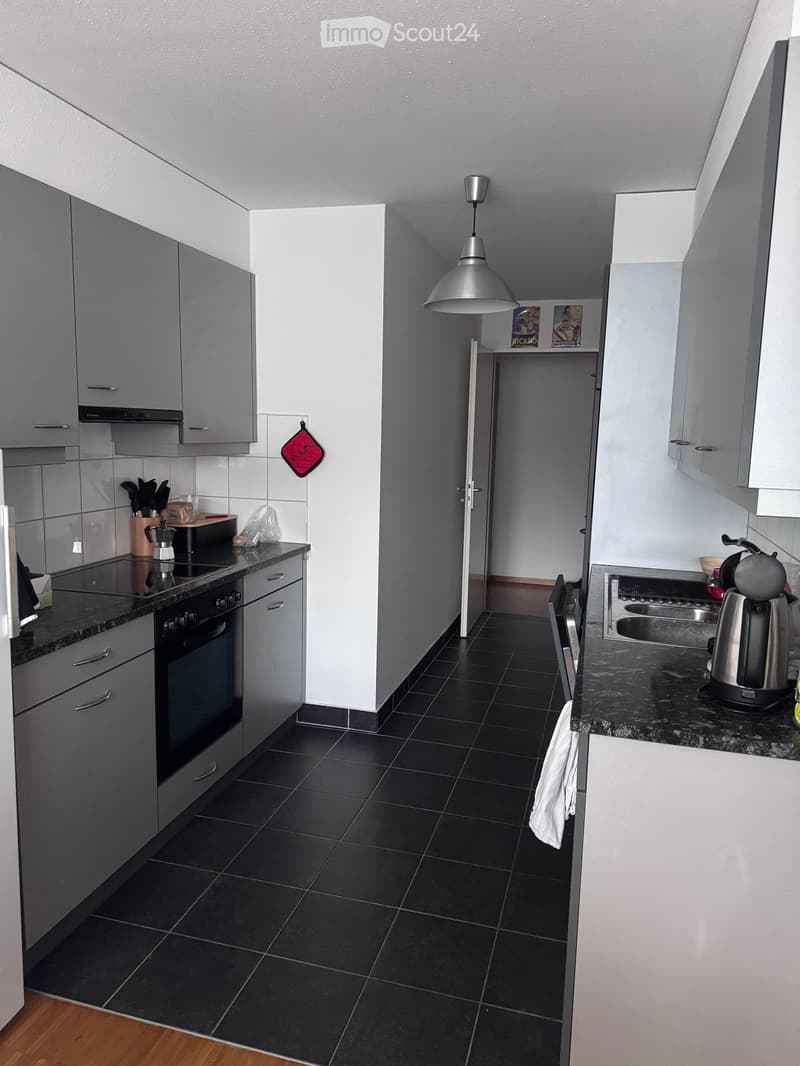 Modern kitchen with grey cabinets, black tiled floor, and stainless steel appliances including an oven, stove, and fridge. A kitchen island with a coffee maker and utensils is visible, along with a hanging pendant light and a small shelf with decorative i
