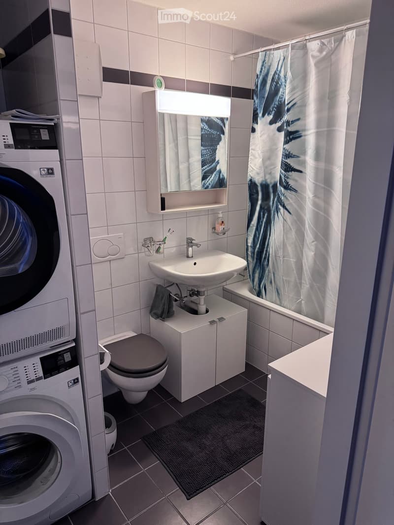 Modern bathroom with white tiles, a sink, mirror, toilet, bathtub with a shower curtain, and a washing machine. Includes a towel and a toothbrush holder.