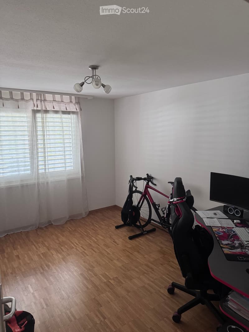 A furnished room with wooden flooring, a desk with a computer and chair, a bicycle on a stand, and a window with curtains.