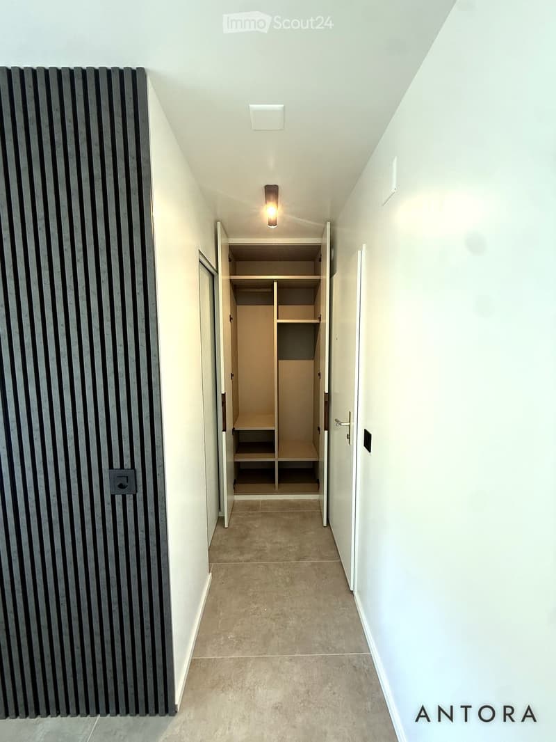 A narrow hallway with white walls, a recessed ceiling light, and an open closet with shelves on the right side.