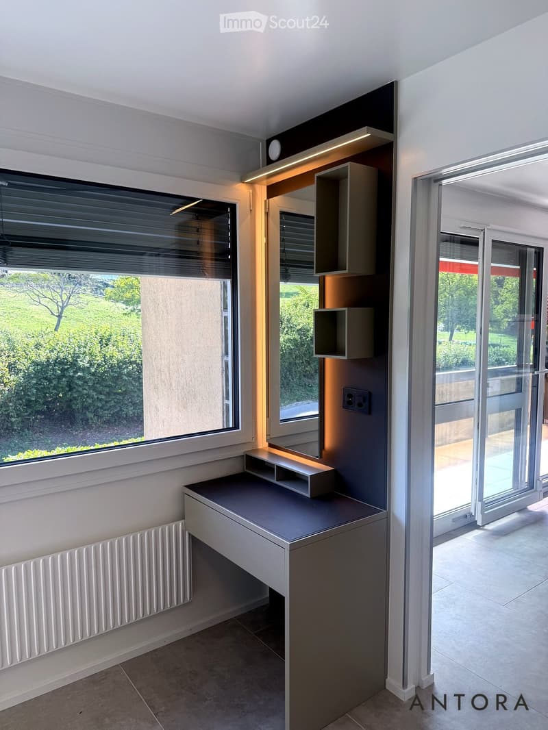 Modern, minimalist room with large windows, built-in shelving, and a sleek desk. The space features a radiator, a sliding glass door leading to an outdoor area, and a clean, contemporary design.