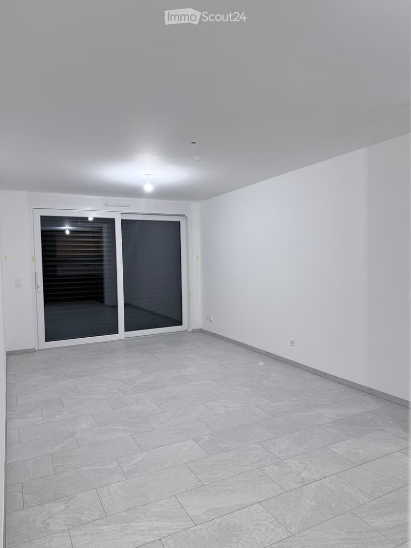 Empty room with white walls, grey tiled floor, and large sliding glass door with blinds.
