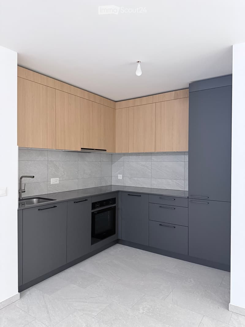 Modern kitchen with light wood upper cabinets, dark grey lower cabinets, grey tiled backsplash, and a stainless steel sink.