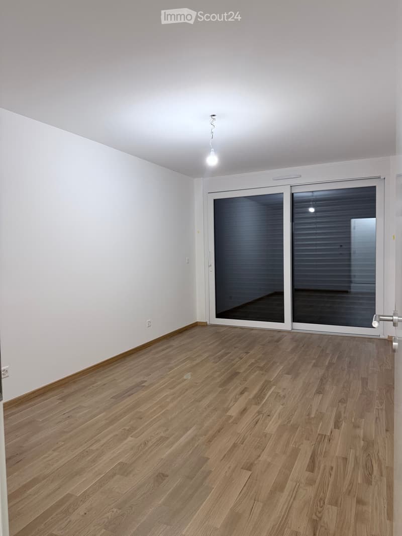 Empty room with light wooden flooring, white walls, and a large sliding glass door with blinds.