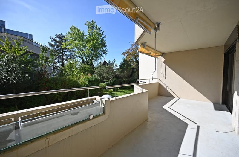 Balcony with modern glass railing, concrete floor, and a retractable awning, overlooking a green garden area.