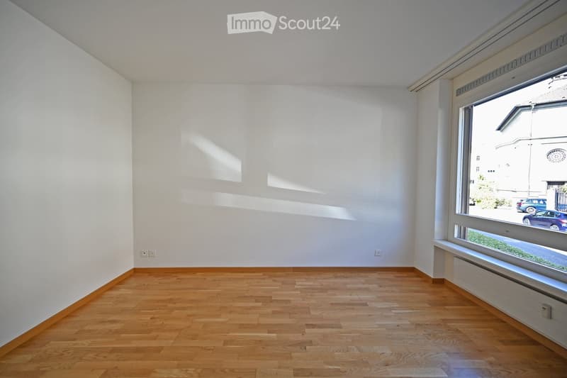 Empty room with light wooden flooring and white walls, featuring a large window with a view of the street and parked cars.